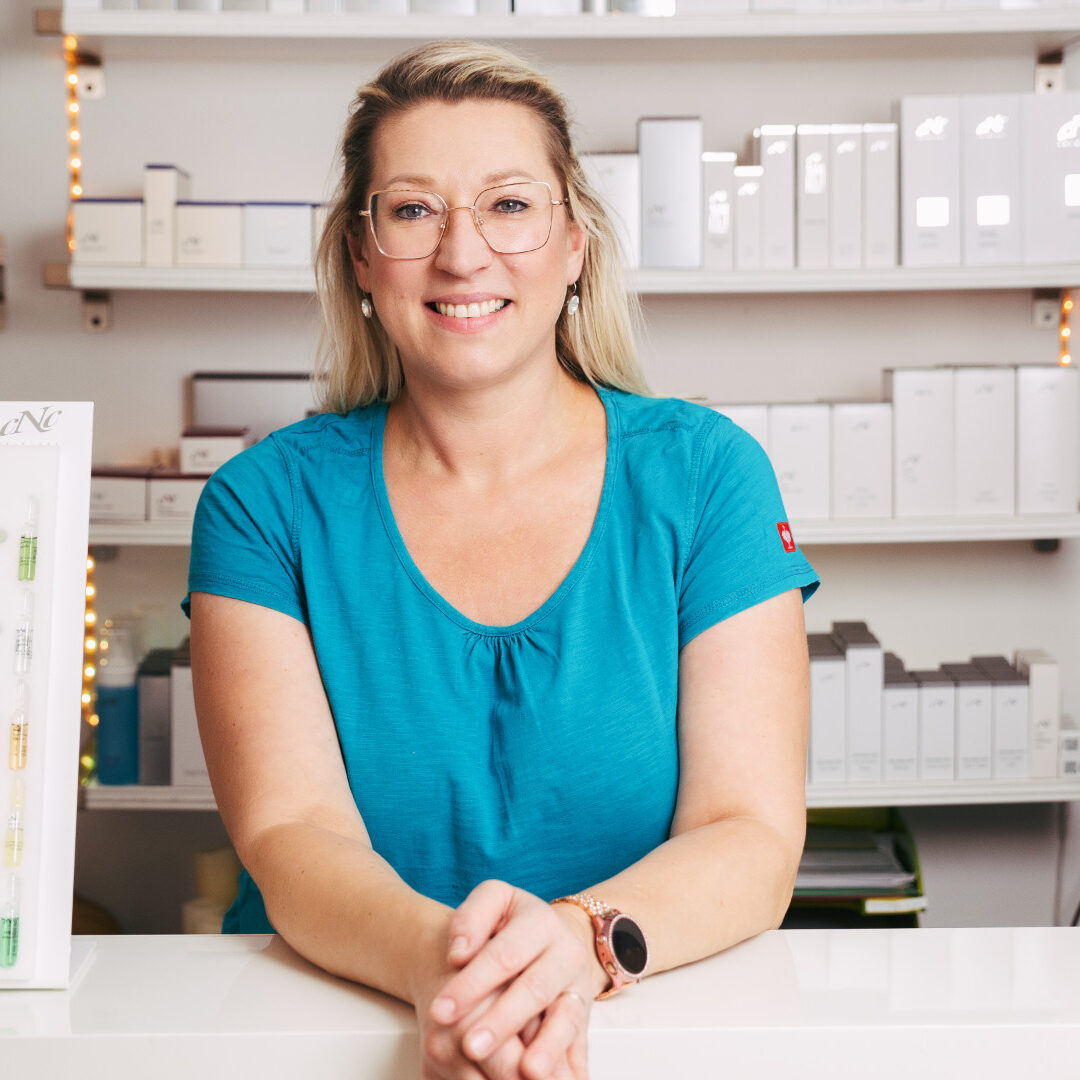 Monika Badauta stützt sich lächelnd auf Theke ab, im Hintergrund befindet sich das Produktregal mit cNc Produkten