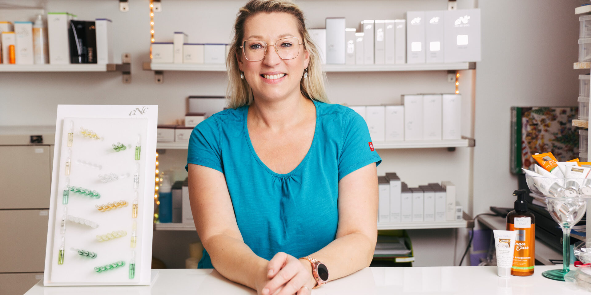 Monika Badauta stützt sich lächelnd auf Theke ab, im Hintergrund befindet sich das Produktregal mit cNc Produkten