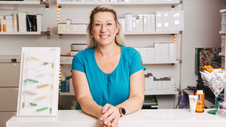 Monika Badauta stützt sich lächelnd auf Theke ab, im Hintergrund befindet sich das Produktregal mit cNc Produkten